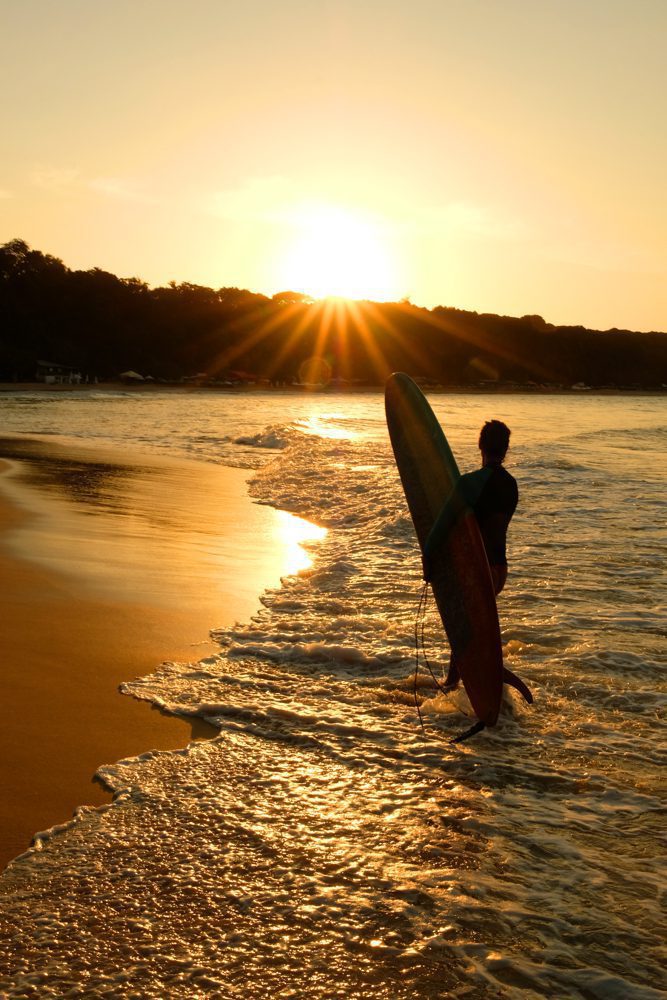 日落时分，男子拿着冲浪板站在Pipa海滩上
